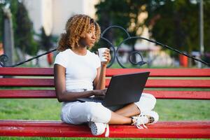 bonita africano americano menina sentado em Banco e falando em celular enquanto trabalhando em computador portátil sentado às mesa com café em rua foto