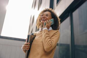 bonita fêmea empreendedor é falando telefone enquanto em pé com computador portátil em moderno construção fundo foto