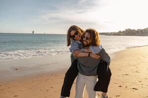 sorridente romântico casal dentro amor abraçando enquanto caminhando ao longo a de praia em ensolarado dia foto