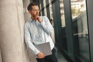 bonito vendas Gerente com computador portátil em pé em moderno coworking fundo e olhando às lado foto