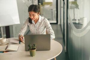 sorridente mulher escritório Gerente fazer notas enquanto trabalhando em computador portátil dentro moderno coworking foto