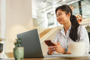 sorridente mulher empreendedor segurando telefone e olhando às lado enquanto trabalhando em computador portátil dentro escritório foto