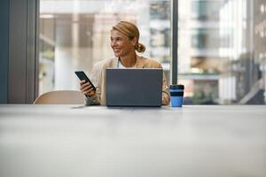 sorridente fêmea empreendedor usar telefone enquanto trabalhando em computador portátil dentro moderno escritório fundo foto