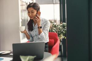 sorridente empresária falando telefone enquanto trabalhando em computador portátil e bebendo café dentro moderno escritório foto