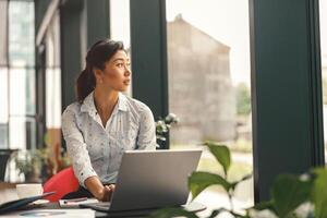 fêmea vendas Gerente trabalhando em computador portátil enquanto sentado dentro moderno coworking e olhando às janela foto