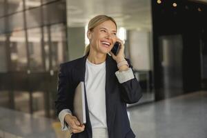 alegre empresária é falando telefone em pé em fundo do moderno escritório corredor e parece longe foto