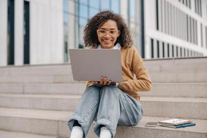 sorridente mulher Gerente trabalhando computador portátil sentado em escadas em moderno construção fundo foto