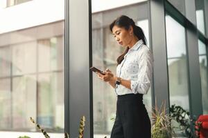 focado o negócio mulher olhando em Smartphone enquanto em pé dentro moderno escritório perto janela foto