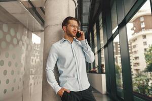 bonito masculino empreendedor falando telefone com cliente enquanto em pé dentro escritório e olhando às lado foto