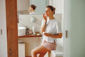 feliz mulher aplicando hidratante hidratante em dela face sentado dentro banheiro. casa beleza rotina foto