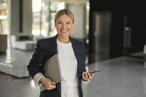 alegre empresária com telefone em pé em fundo do moderno escritório corredor e parece Câmera foto