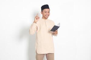 retrato do atraente ásia muçulmano homem dentro Koko camisa com peci lendo uma livro, dizendo este ele tem a idéia enquanto apontando dedo e caneta. isolado imagem em branco fundo foto