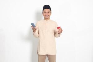 retrato do atraente ásia muçulmano homem dentro Koko camisa com calota craniana segurando uma Móvel telefone e apresentando crédito cartão. isolado imagem em branco fundo foto