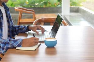 fechar acima do masculino mão escrevendo em Nota livro com caneta, mostrando em branco tela do computador portátil e Smartphone. trabalhando dentro cafeteria conceito com uma copo do café foto