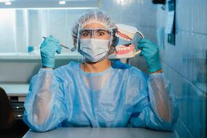 uma dental médico vestindo azul luvas e uma mascarar detém uma dental modelo do a superior e mais baixo mandíbulas e uma dental espelho foto