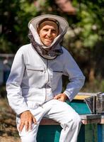 bonito adulto homem apicultor dentro branco protetora terno às apiário. verão jardim com urticária borrado fundo. fechar-se. foto