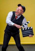 Senior bonito homem segurando uma cinema badalo. homem vestindo terno com não jaqueta. pessoa isolado contra amarelo fundo. foto