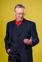 retrato do Senior homem de negocios segurando dentro mãos telefone enquanto em pé às estúdio sobre amarelo fundo. o negócio pessoas conceito. foto