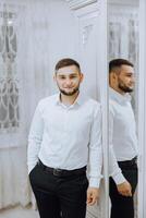 retrato do uma jovem homem dentro uma branco camisa e luz calça dentro uma sala. a noivo é preparando para a Casamento foto