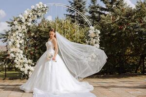 uma lindo noiva dentro uma exuberante branco Casamento vestir contra a fundo do uma cerimonial arco. foto
