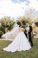 à moda, jovem noivo e lindo noiva dentro uma grandes branco vestir e uma grandes véu com uma ramalhete dentro seus mãos, abraçando dentro a parque dentro a outono natureza. Casamento retrato do recém-casados. foto