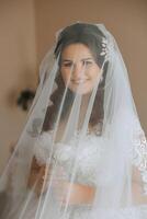 lindo jovem noiva segurando véu dentro branco Casamento vestir, retrato do morena noiva dentro hotel sala, manhã antes casamento. foto