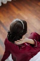 manhã retrato do uma luxuoso e lindo noiva dentro uma hotel dentro uma luxuoso quarto dentro vermelho pijamas, sentado a partir de a costas e mostrando fora dela Penteado. foto