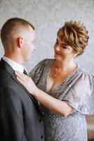 mãe ajuda dela adulto filho preparar para a Casamento cerimônia. a emocional e tocante momento às uma casamento. uma mãe abraços dela filho foto