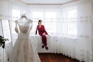 manhã retrato do uma luxuoso e lindo hotel noiva dentro uma luxo suíte dentro vermelho pijamas sentado em a peitoril da janela Próximo para dela Casamento vestir. foto