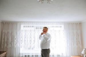 uma homem dentro uma branco camisa carrinhos de a janela dentro a quarto e aperta a botões em dele colarinho e mangas. Assistir em mão. à moda o negócio retrato do uma homem, fechar-se foto. a noivo é preparando. foto