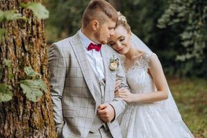 adorável e à moda recém-casados estão abraçando e sorridente contra a fundo do outono natureza dentro uma lindo jardim. a incrivelmente lindo jovem noiva inclinou-se contra a ombro do dela Amado noivo foto