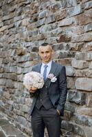 retrato do uma bonito jovem noivo em a outono dia ao ar livre dentro a elegante terno e segurando uma Casamento ramalhete do flores ao ar livre foto. bonito homem dentro uma o negócio terno. foto