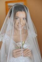 lindo jovem noiva segurando véu dentro branco Casamento vestir, retrato do morena noiva dentro hotel sala, manhã antes casamento. foto