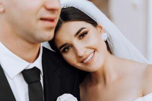 retrato do uma sorridente noiva com uma ramalhete do flores abraçando dela marido enquanto olhando às a Câmera. lindo Casamento celebração. Casamento Penteado com uma véu. casal dentro amar. foto