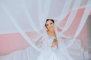 lindo jovem noiva segurando véu dentro branco Casamento vestir, retrato do morena noiva dentro hotel sala, manhã antes casamento. foto