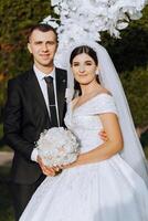 à moda, jovem noivo e lindo noiva dentro uma grandes branco vestir e uma grandes véu com uma ramalhete dentro seus mãos, abraçando dentro a parque dentro a outono natureza. Casamento retrato do recém-casados. foto