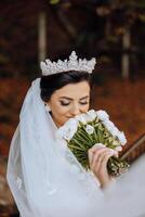 retrato. uma morena noiva dentro uma véu e uma chique coroa poses e cheira dela ramalhete. prata joia. lindo Maquiagem e cabelo. outono casamento. celebração foto