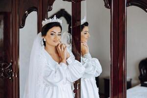 dentro a manhã, a incrivelmente lindo e luxuoso noiva, vestido dentro uma branco manto, poses de a espelho dentro dela quarto. manhã do a noiva com uma luxuoso Penteado e natural inventar. foto