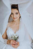 lindo jovem noiva segurando véu dentro branco Casamento vestir, retrato do morena noiva dentro hotel sala, manhã antes casamento. foto