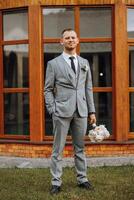 retrato do uma bonito jovem noivo em a outono dia ao ar livre dentro a elegante terno e segurando uma Casamento ramalhete do flores ao ar livre foto. bonito homem dentro uma o negócio terno. foto