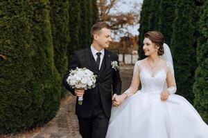 uma Casamento casal é caminhando dentro natureza em a outono dia. feliz jovem noiva e elegante noivo segurando mãos. uma à moda casal do recém-casados em seus Casamento dia. foto