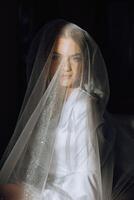 retrato do a incrivelmente lindo menina noiva dentro uma branco roupão dentro a quarto, a noiva poses segurando dela véu dentro dela mãos e cobre ela mesma com isto. foto