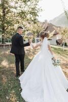 Casamento retrato. uma noivo dentro uma Preto terno e uma Loiras noiva andar segurando mãos dentro uma jardim. traseiro visualizar. uma branco, grandes véu dentro a ar. foto sessão dentro natureza. lindo cabelo e Maquiagem