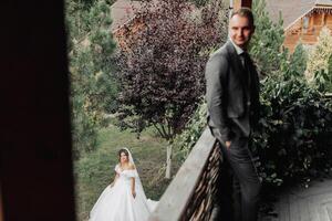 Casamento foto. a noiva dentro uma volumoso branco vestir e uma grandes véu anda em dentro a jardim ao longo uma pedra caminho, a noivo carrinhos em a sacada dentro a primeiro plano, dele ombros virou para dela. foto