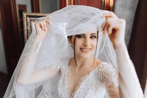 lindo jovem noiva segurando véu dentro branco Casamento vestir, retrato do morena noiva dentro hotel sala, manhã antes casamento. foto