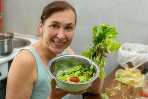 uma mulher mostra uma tigela do legumes e ervas com alface folhas contra a fundo do a cozinha. foto