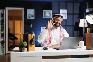 muçulmano homem acenando às dele pessoal computador enquanto sentado às uma escrivaninha tendo uma vídeo chamar. ele usa dele digital computador portátil para explorar, bater papo, e Faz pesquisa enquanto imerso dentro digital tecnologia. foto