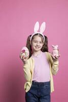 alegre criança pequena apresentando uma Rosa ovo e uma recheado Coelho dentro estúdio, vestindo tranças e fofa Coelho orelhas em Câmera. sorridente jovem criança mostrando dela Páscoa decorações e colorida brinquedos. foto