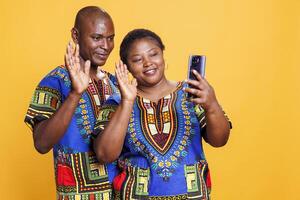 sorridente africano americano homem e mulher vestindo étnico roupas acenando Oi enquanto Falando dentro Smartphone Video chamada. alegre casal mostrando cumprimento gesto enquanto tendo conectados comunicação em telefone foto