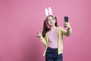 alegre criança pequena usa Smartphone aplicativo para uma Video chamada bate-papo contra Rosa fundo, alegre pequeno criança dentro encontro com dela amigos em conectados Videoconferência. sorridente positivo menina com Coelho ouvidos. foto